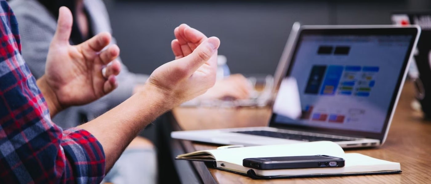 Two Individuals sitting at a table with a laptop and one person making hand gestures to communicate.